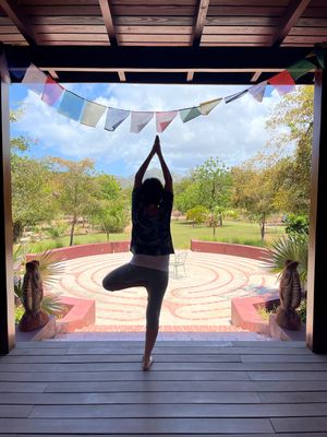 Person performing tree pose yoga on a wooden deck with colorful prayer flags and a garden labyrinth in background.