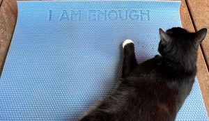 Black cat lying on a light blue yoga mat with the text I AM ENOUGH embossed on it.