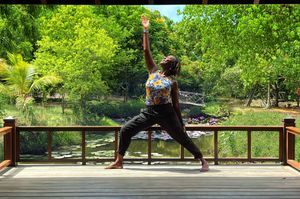 Person performing warrior yoga pose on wooden deck surrounded by lush green trees and serene pond backdrop.