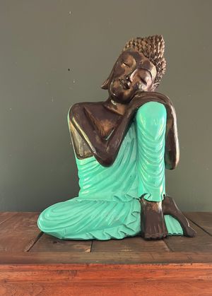 Bronze and turquoise Buddha statue seated in a resting pose on a wooden surface against a green wall.