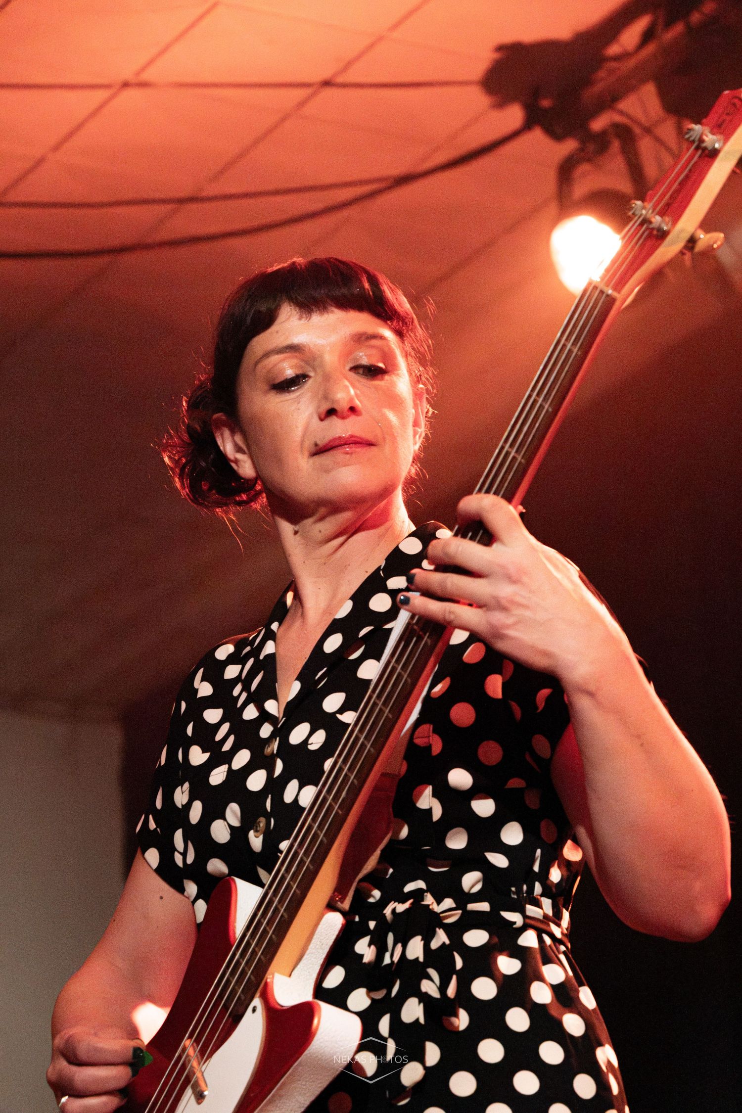 Woman in polka dot dress playing bass guitar on stage under red and white stage lighting.