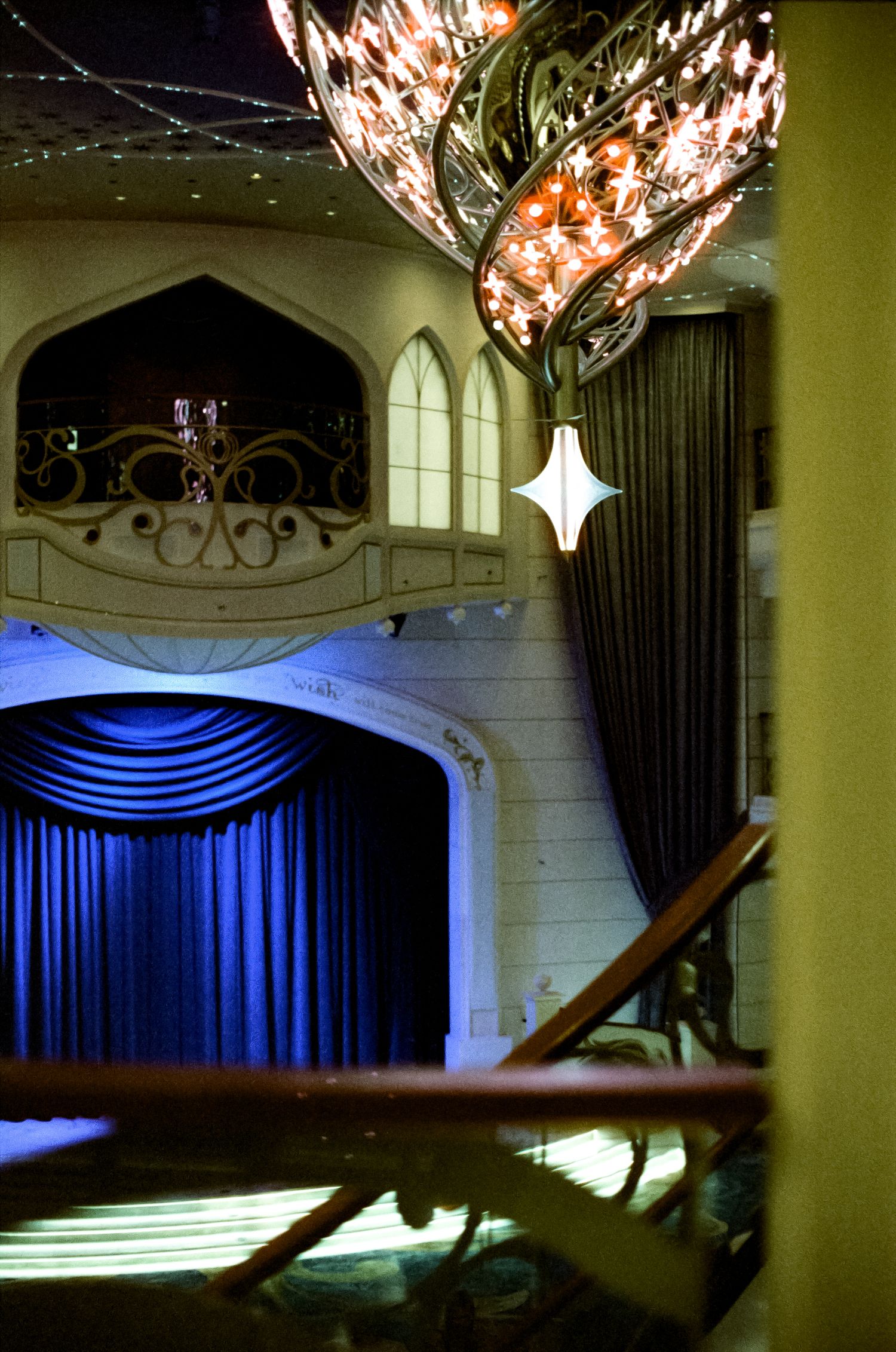 Elegant theater interior with blue velvet stage curtains, ornate chandelier, and Gothic-arched balcony in warm lighting.