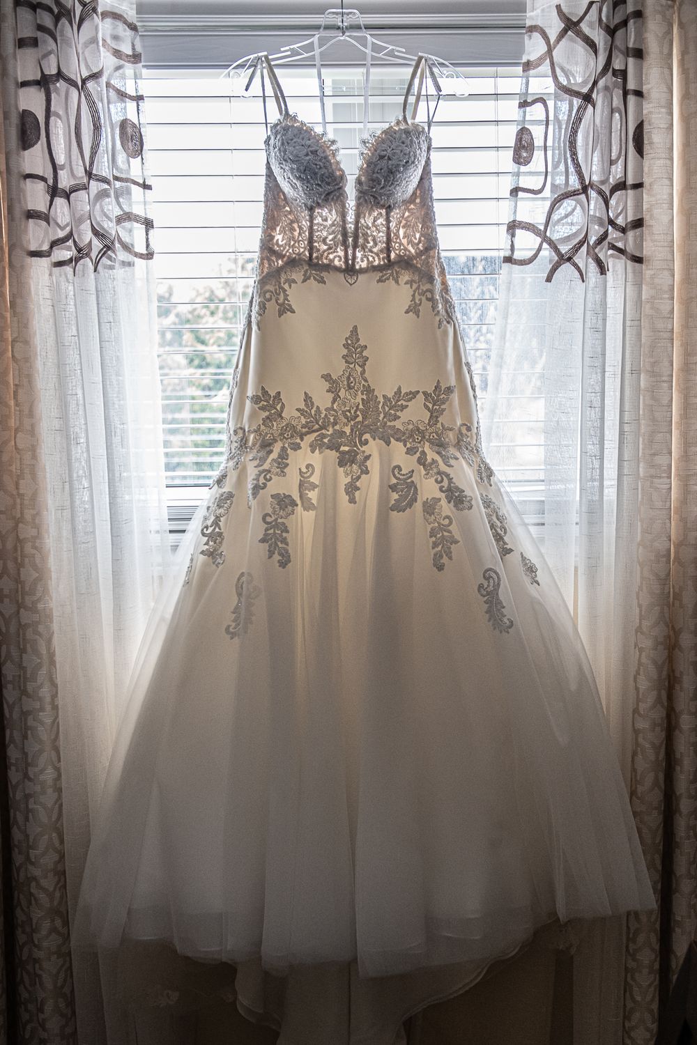 Elegant beaded wedding gown with silver floral embroidery hanging in window light.