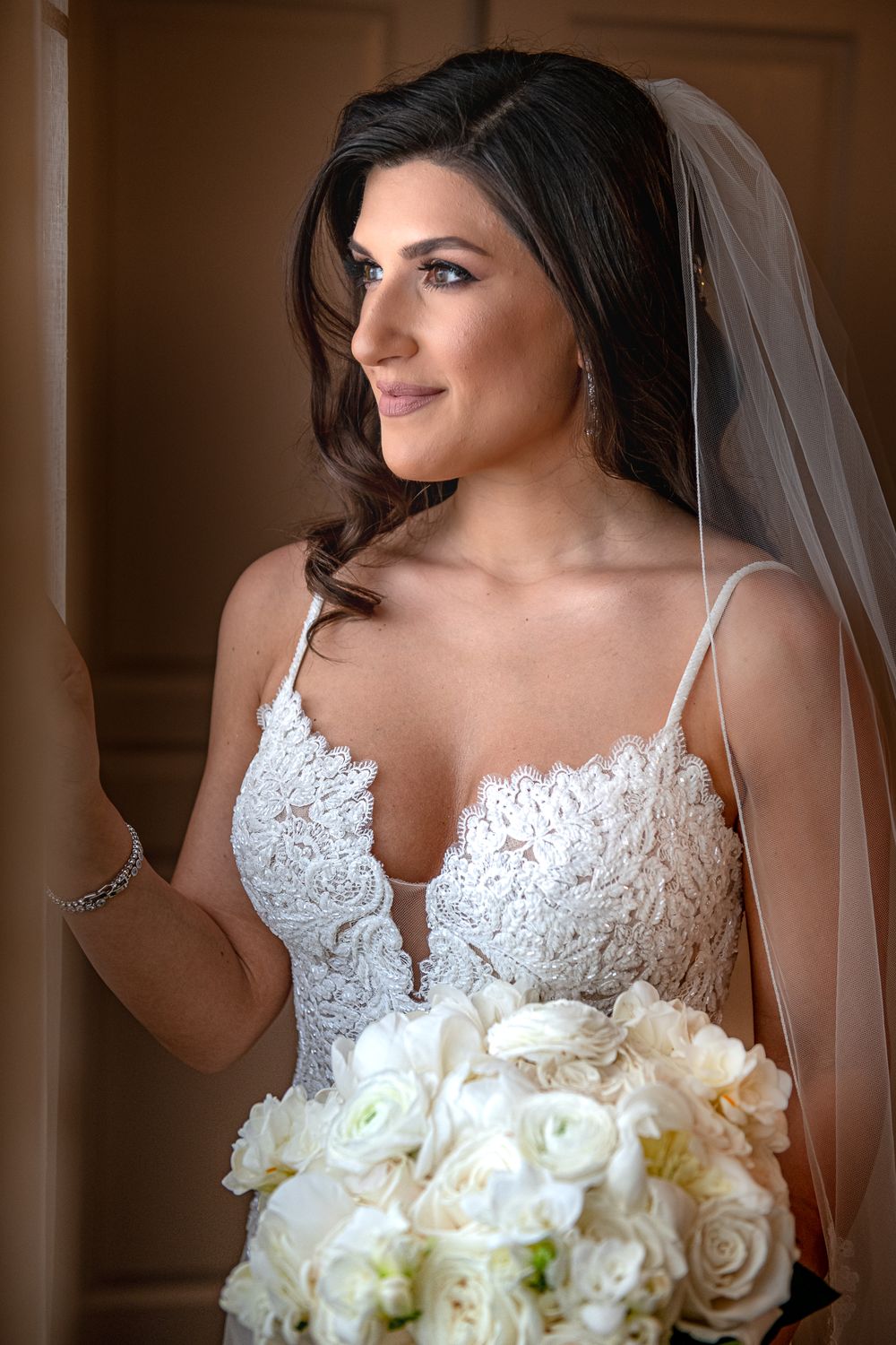 Beautiful bride in lace gown holds white floral bouquet while gazing out a window on her wedding day.