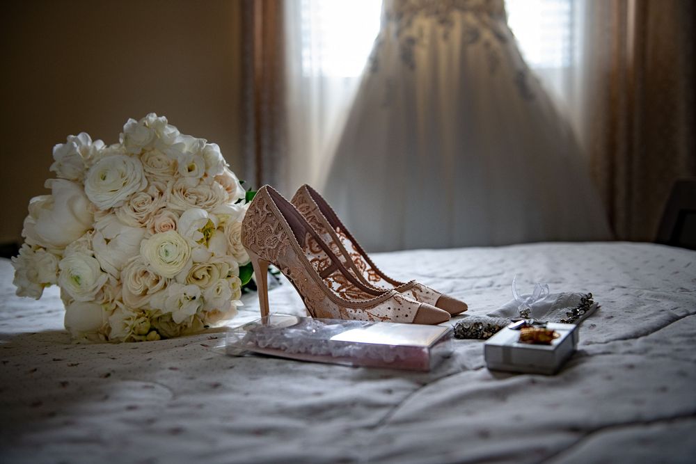 Lace bridal heels beside white rose bouquet and accessories on bed before wedding.