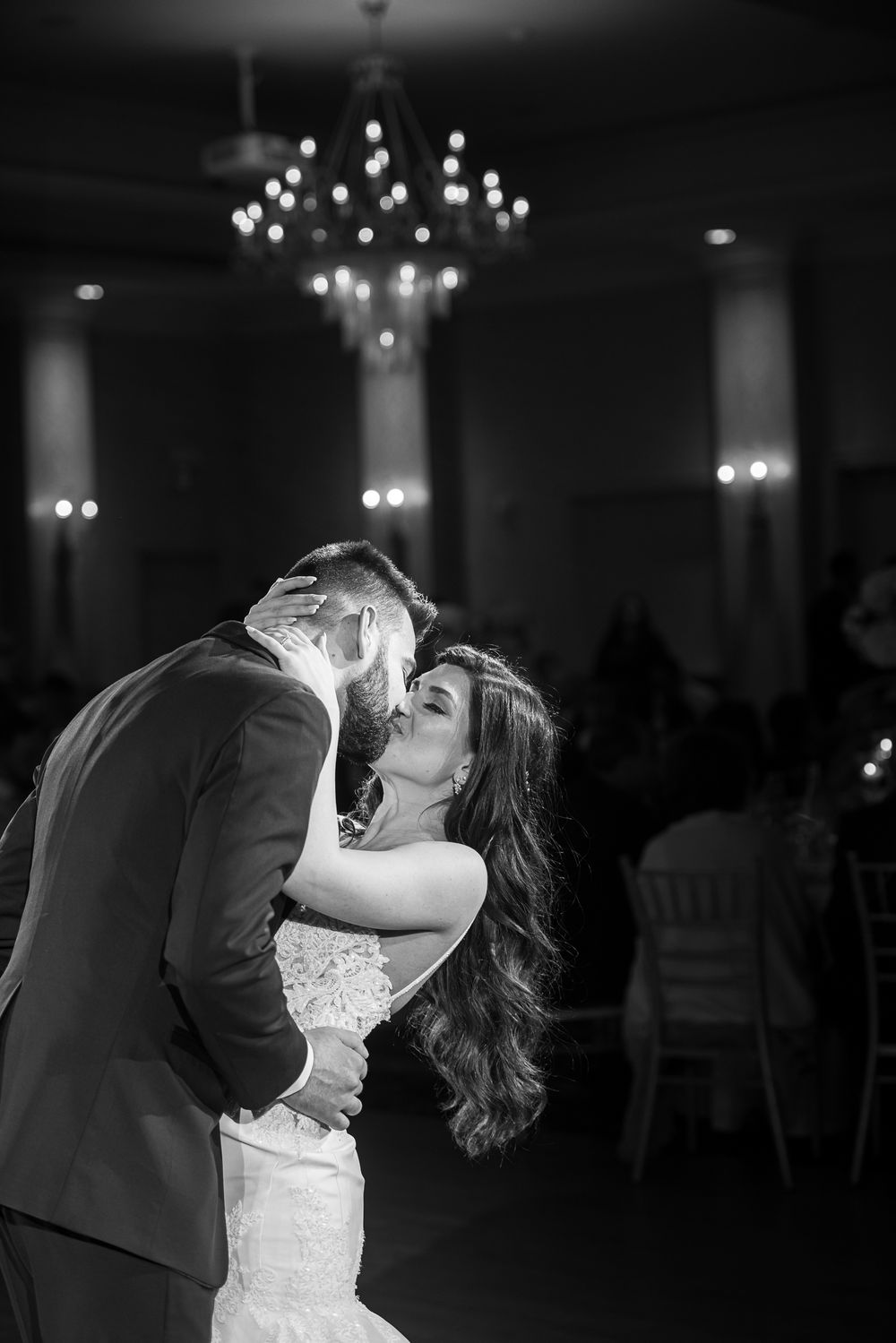 Black and white photo of bride and groom kissing passionately on dance floor beneath chandelier.
