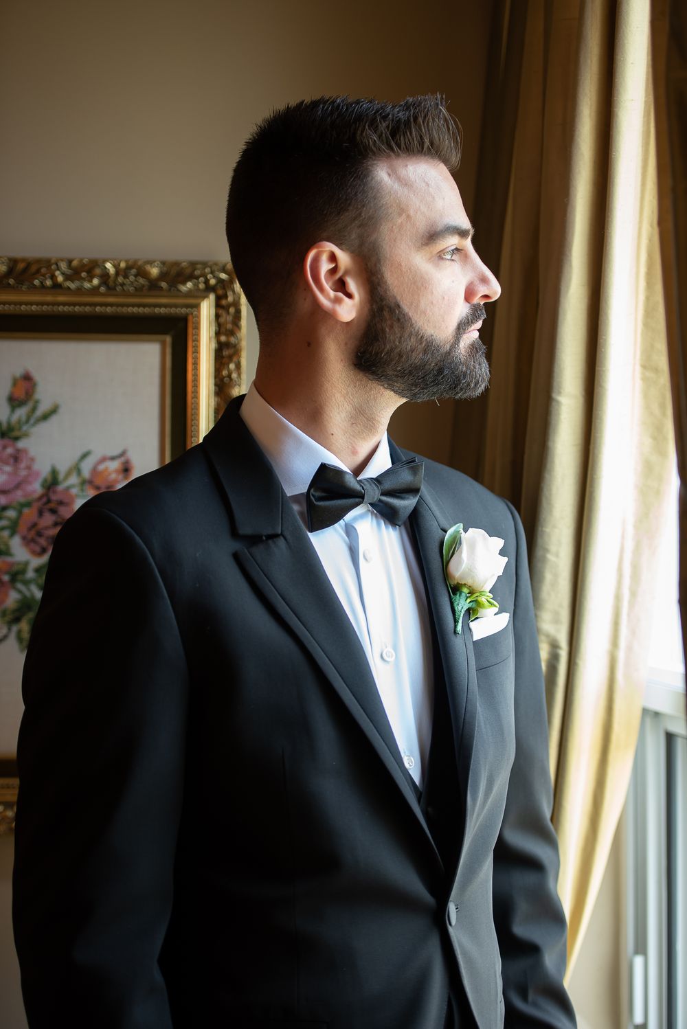 Handsome bearded groom in black tuxedo with bow tie and boutonniere gazing out a window.