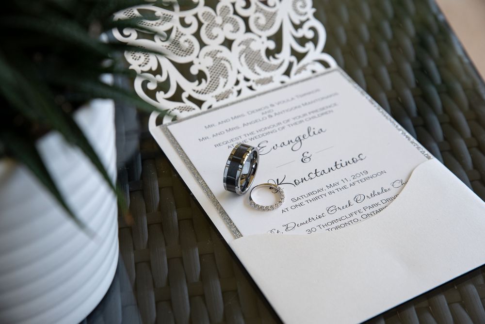Wedding invitation with couple names Evangelia and Konstantinos alongside two rings on decorative surface.