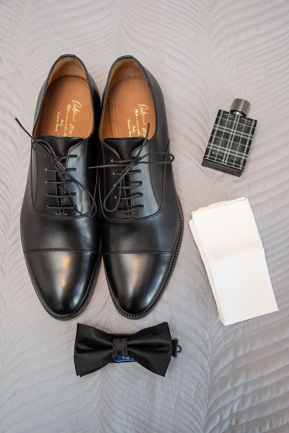 Black leather Oxford dress shoes with bow tie, cologne, and pocket square laid flat on a white surface.