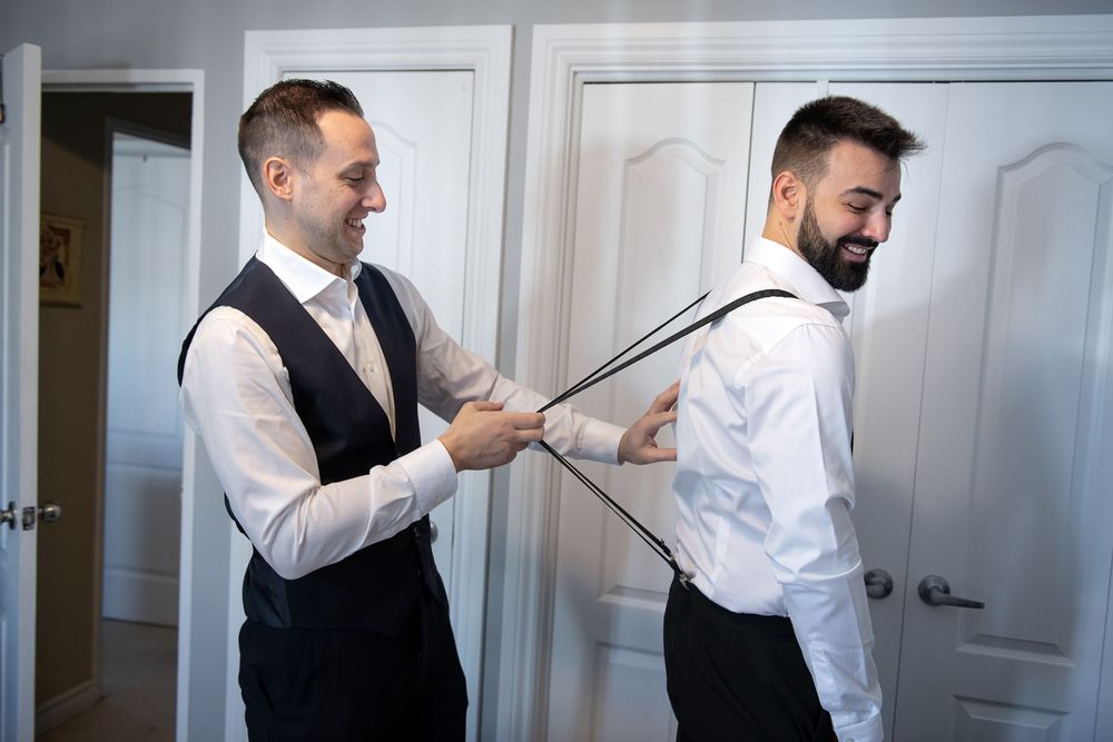 Groomsman helping groom adjust suspenders while both smile during wedding day preparations indoors.