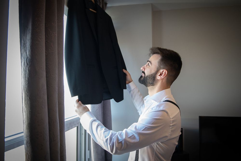 Bearded groom in white shirt and suspenders examining his dark suit jacket hanging by a window.