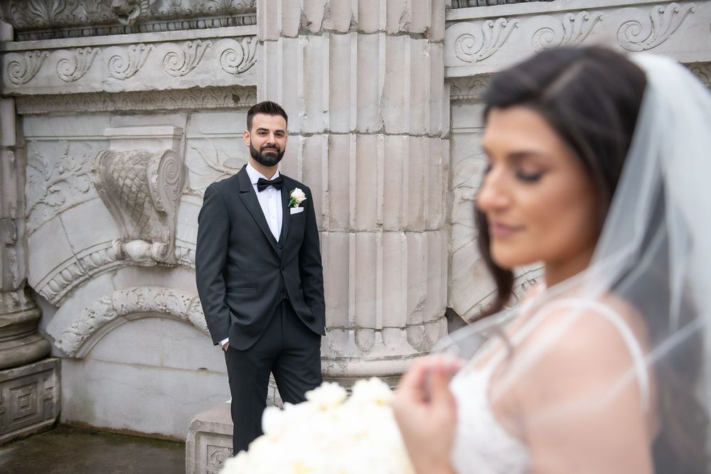 Groom in black tuxedo stands behind bride in soft focus holding white roses at stone architecture venue.