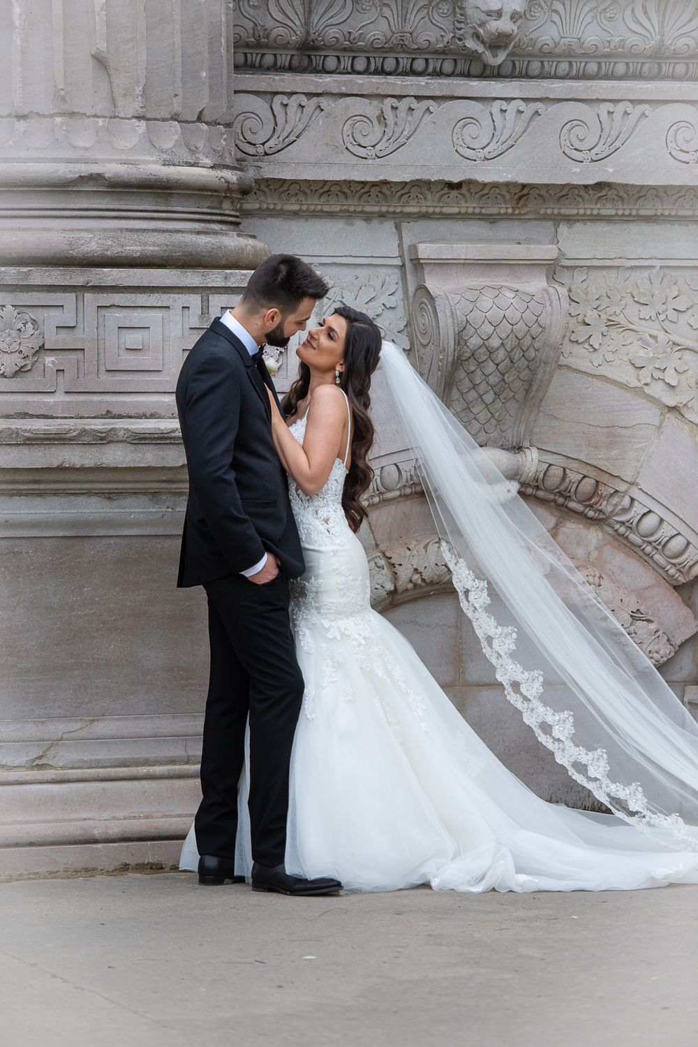 Bride in lace mermaid gown with cathedral veil and groom in black tuxedo sharing intimate moment by carved stone wall.