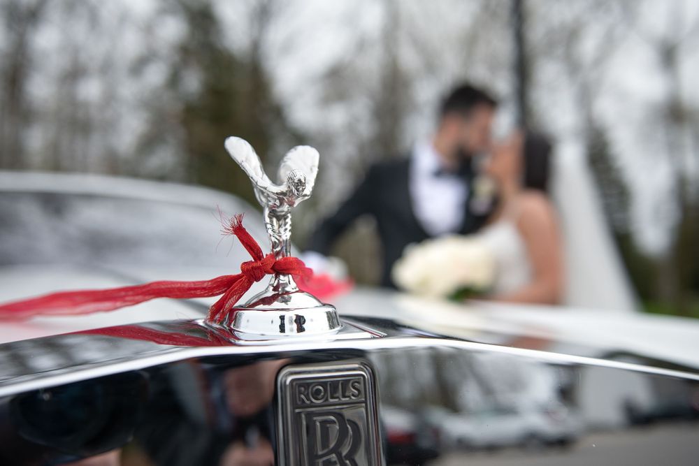 Close-up of Rolls-Royce hood ornament with red ribbon bow and blurred bride and groom kissing in background.