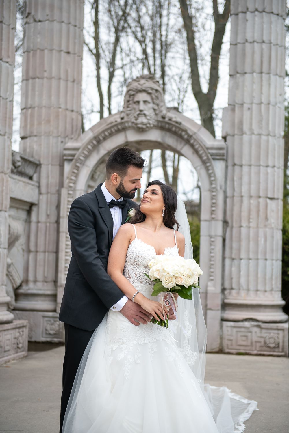 Bride and groom embrace before ornate stone arch with white rose bouquet on their wedding day.
