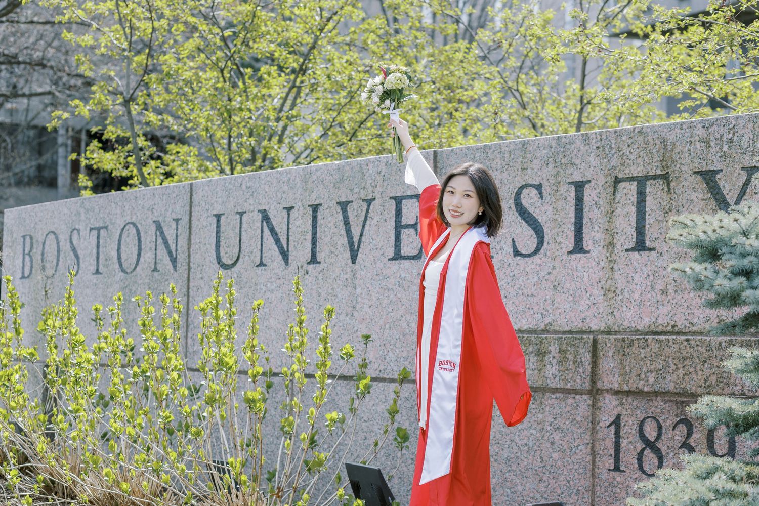 Boston University Graduate in red regalia celebrates commencement in front of Boston University stone sign surrounded by spring foliage.