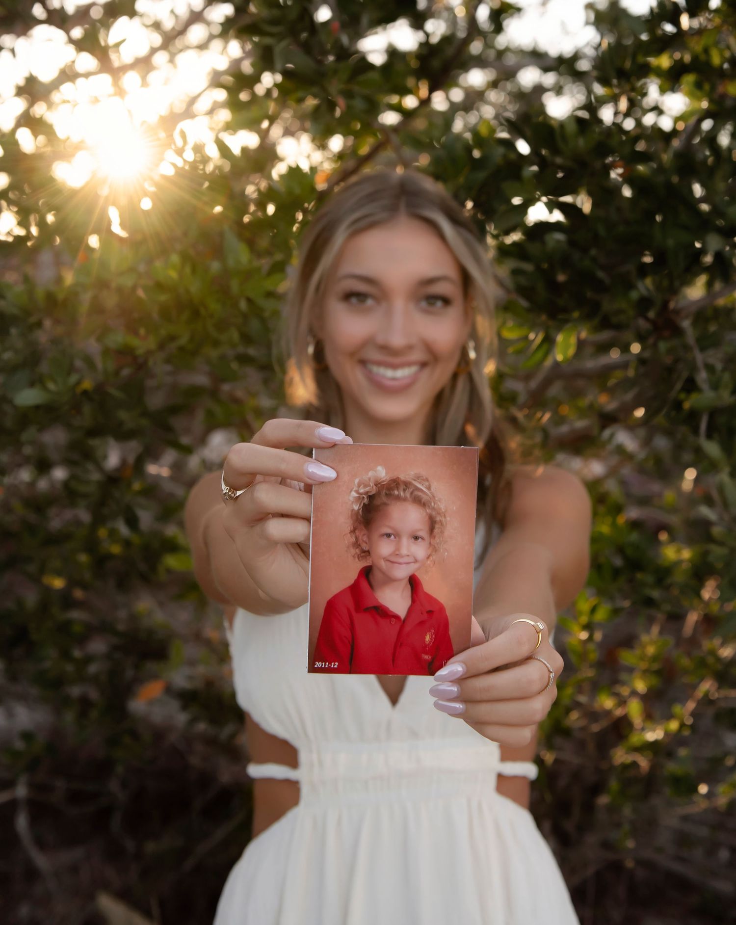 Young blonde woman in white dress holds up her childhood school photo outdoors at golden hour.