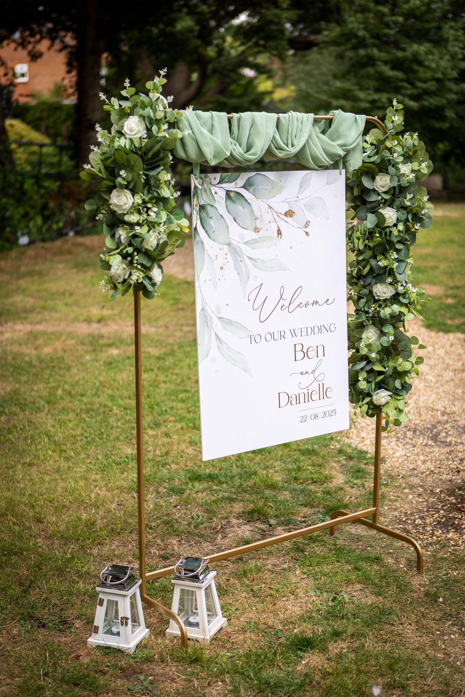 Elegant wedding welcome sign for Ben and Danielle decorated with green foliage and white flowers on a gold stand.