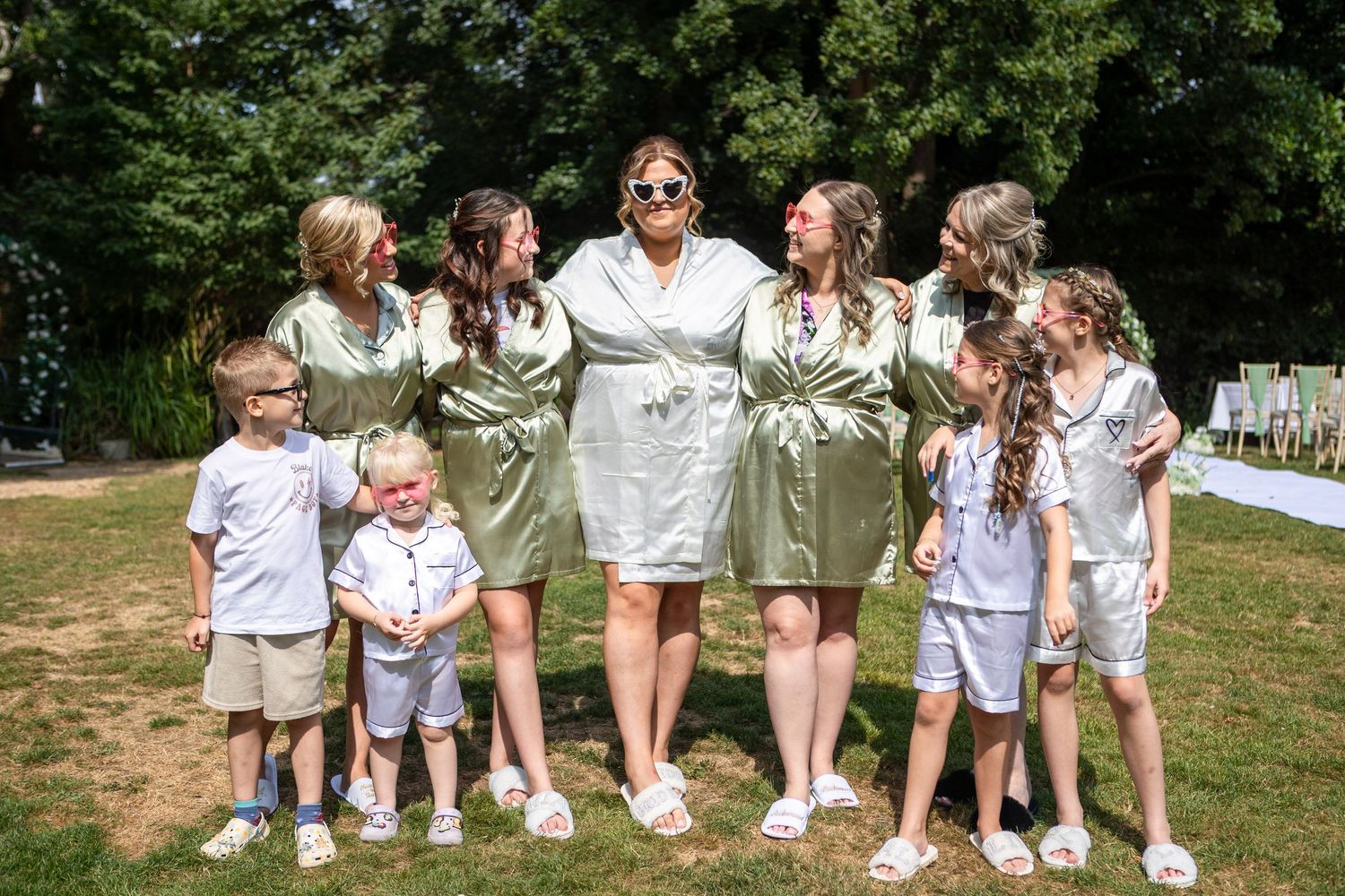Bride in sage green robe posing with bridesmaids and children in matching outfits on a sunny lawn.