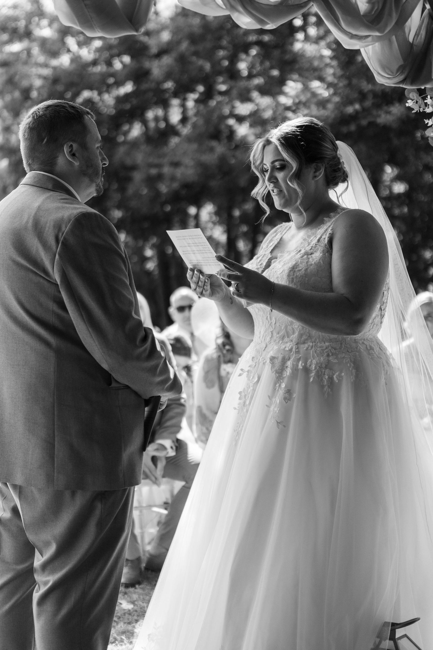 Black and white photo of a bride reading her wedding vows to the groom during an outdoor ceremony.