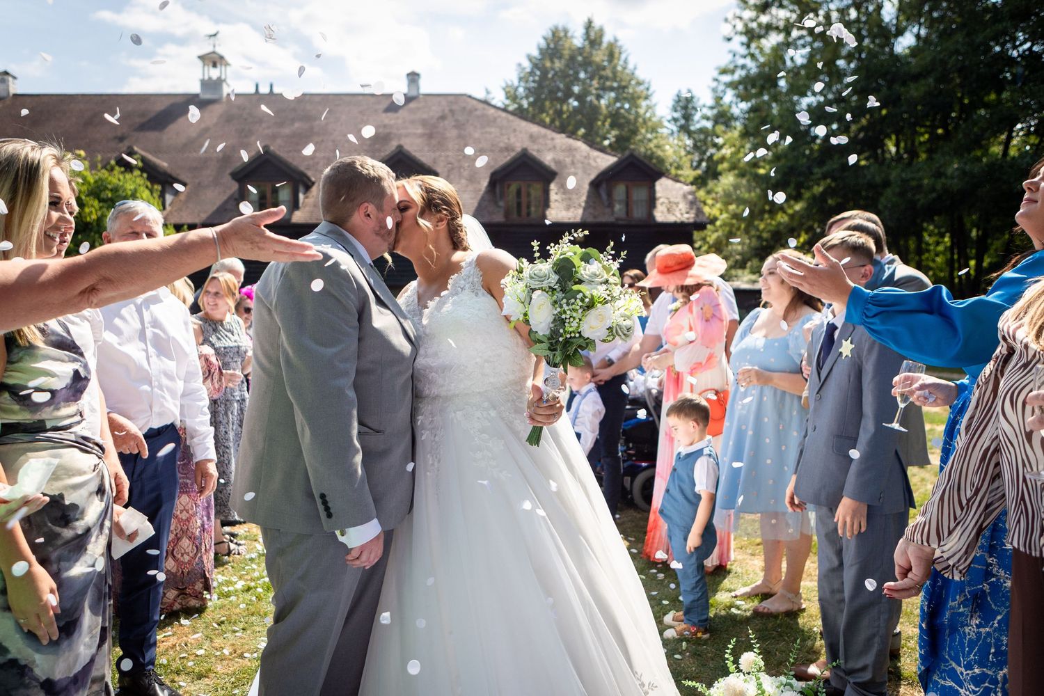 Newlyweds kissing as wedding guests shower them with white confetti outside a rustic venue.