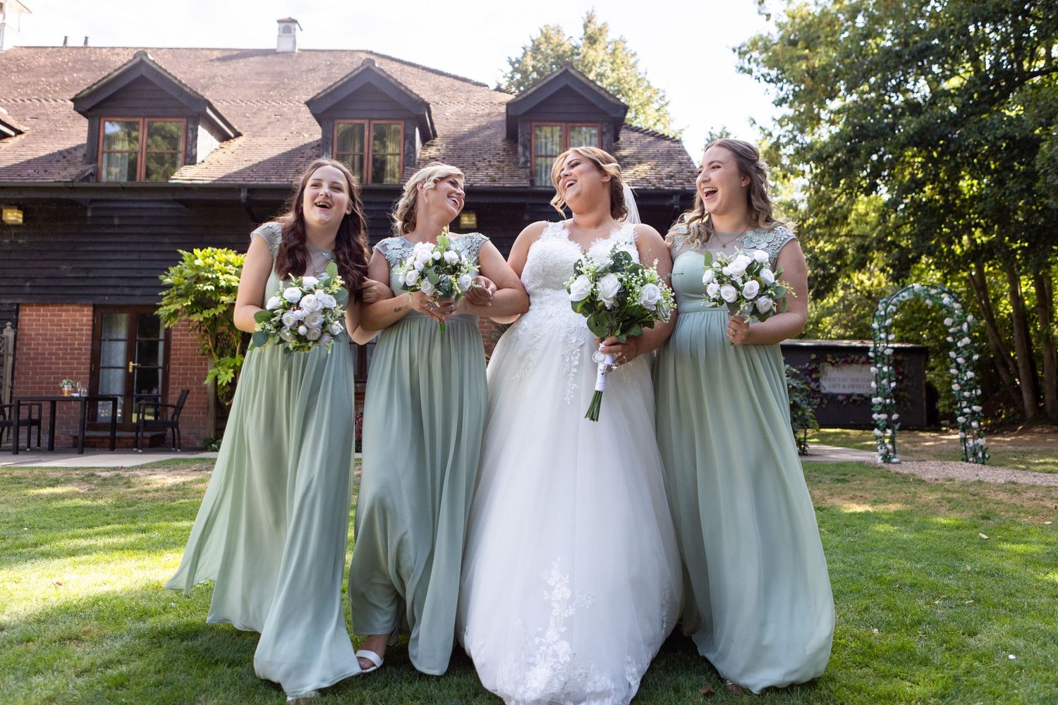 Bride laughing joyfully with three bridesmaids in sage green gowns holding white bouquets outdoors.