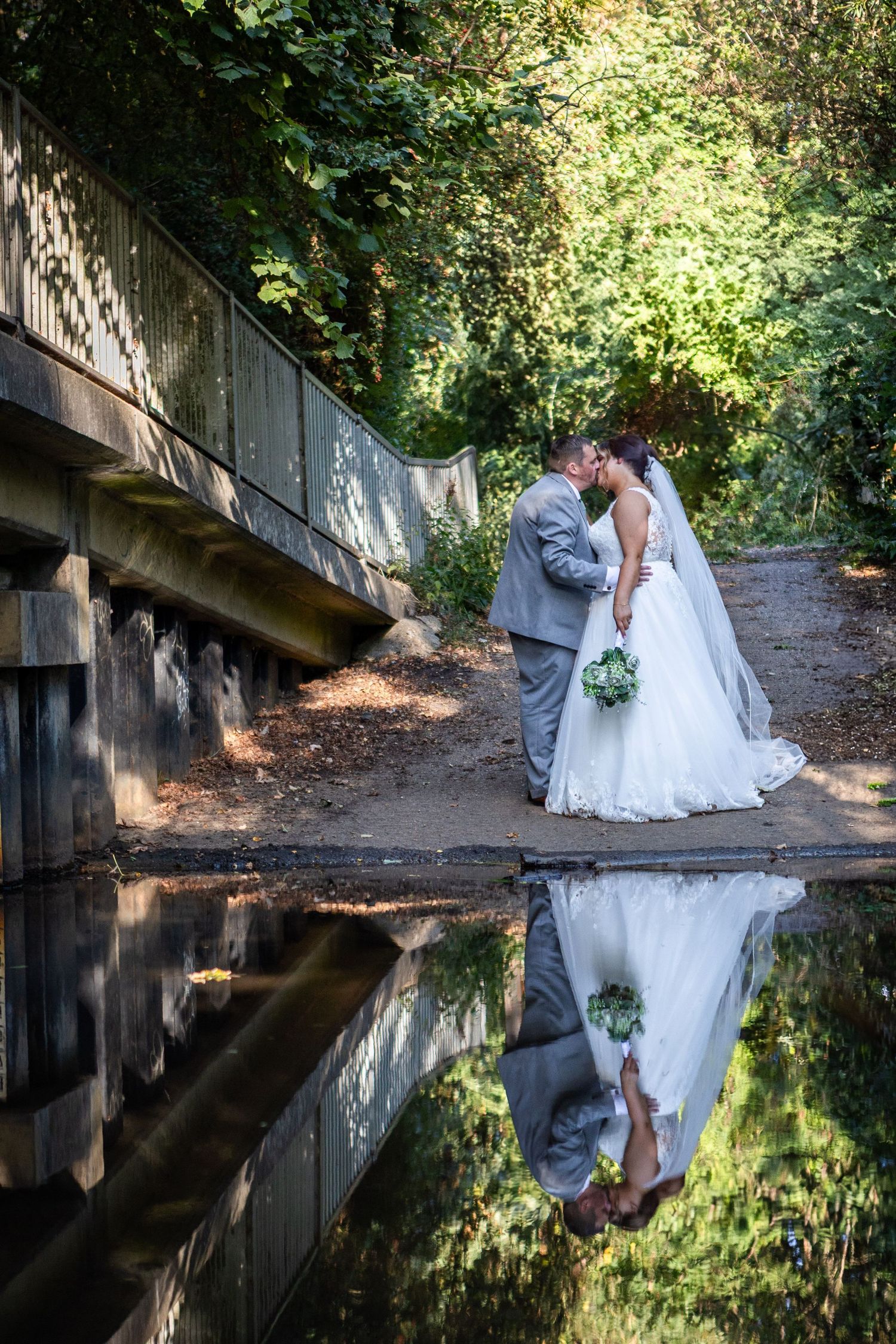 Bride and groom kissing near a bridge with their reflection mirrored in the still water below.