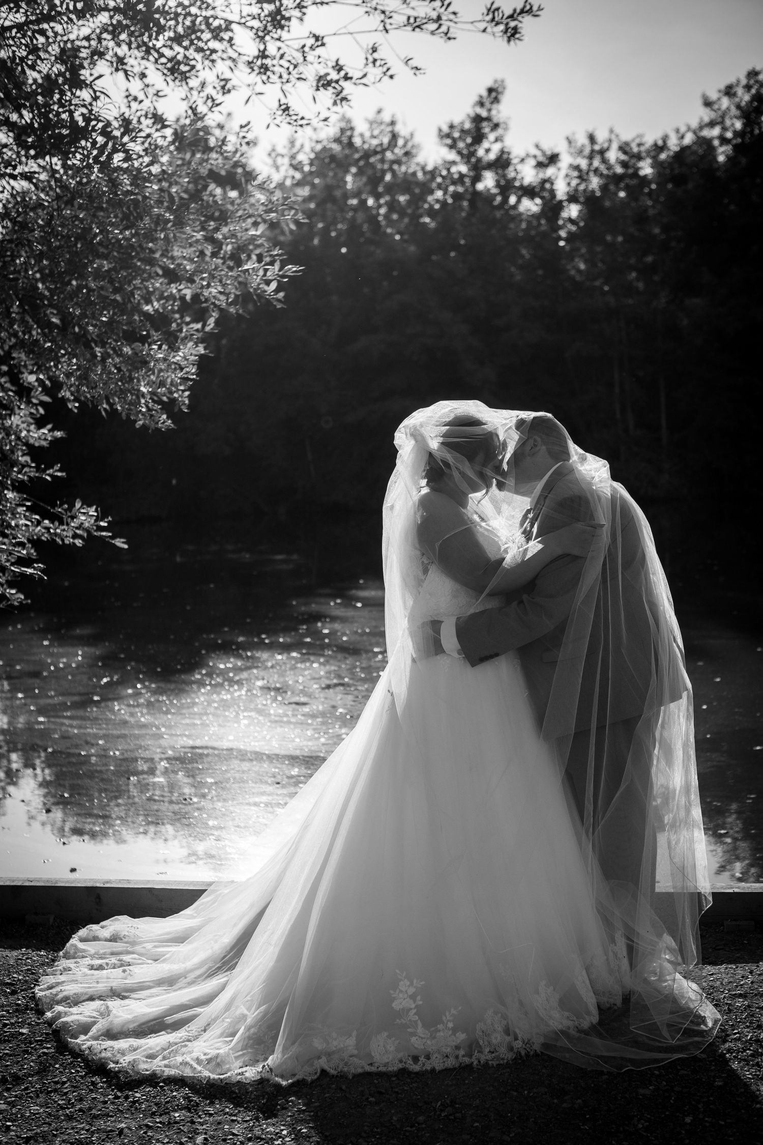 Black and white photo of a bride and groom sharing a kiss by a pond, with her veil draped over them both.