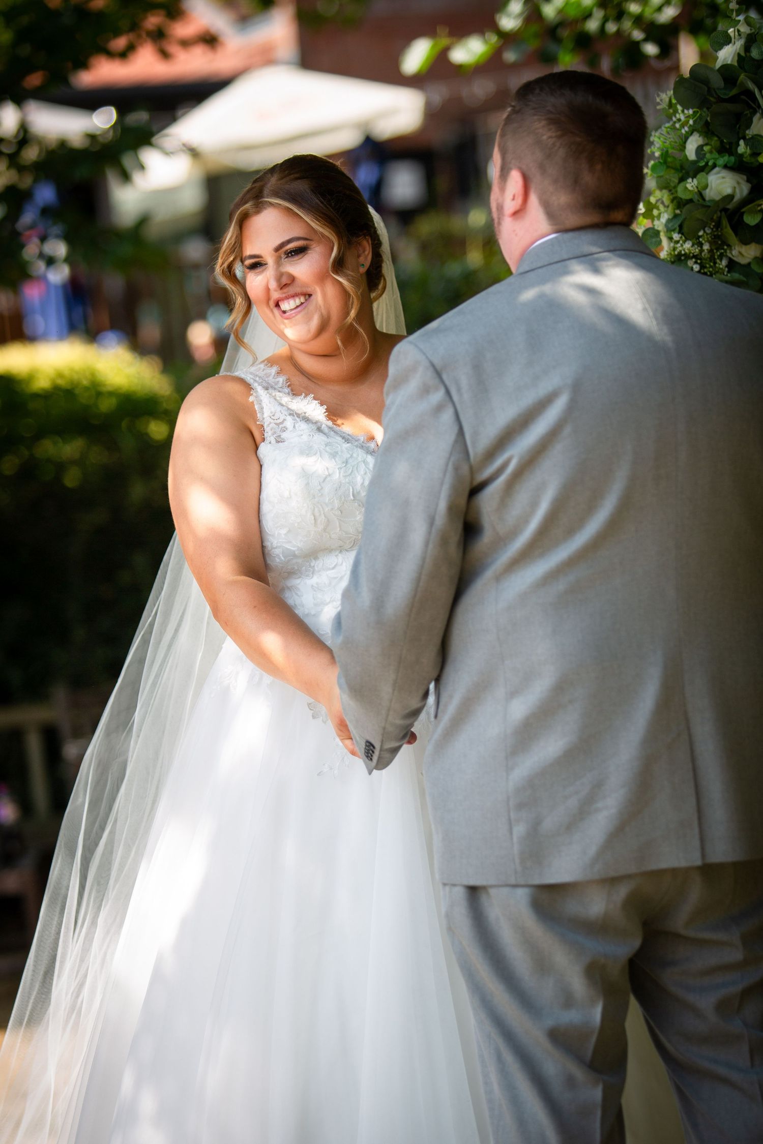 Smiling bride in lace wedding gown holds hands with groom in gray suit during outdoor wedding ceremony.