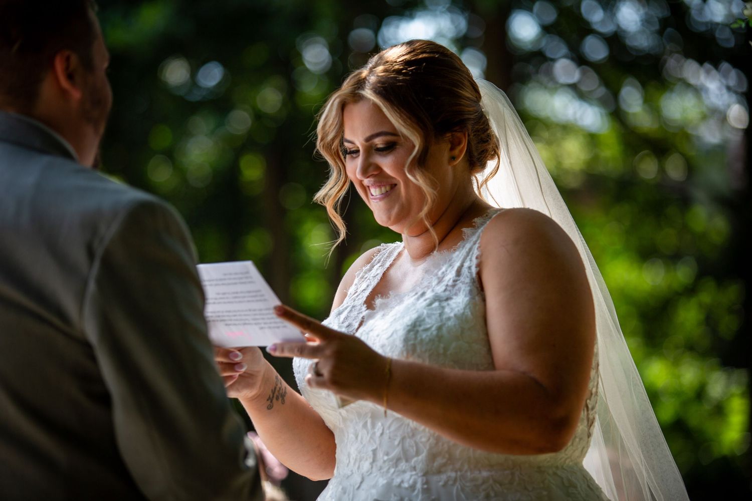 Smiling bride in lace veil reads personal vows from a card during an outdoor garden wedding ceremony.