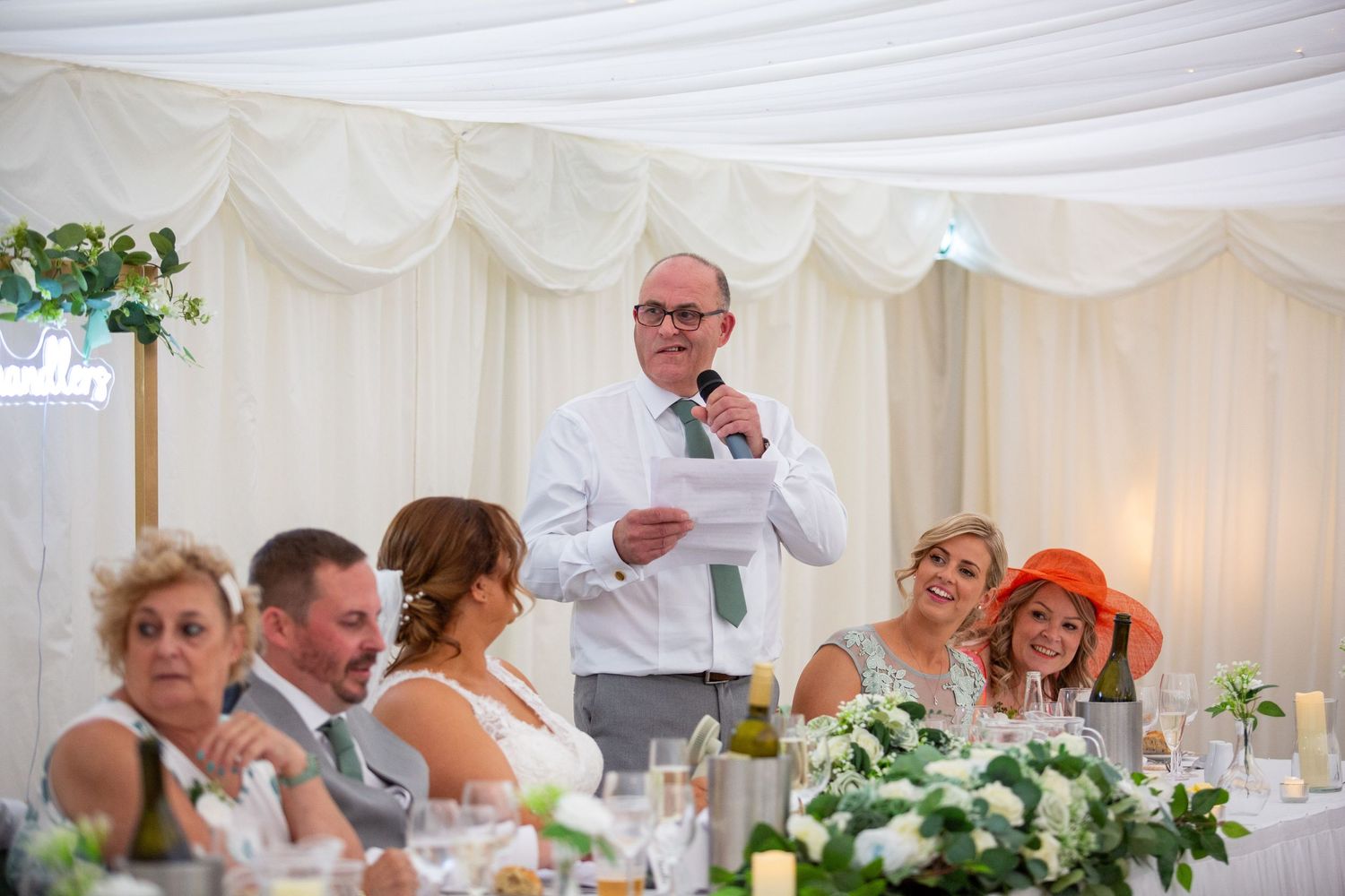 Wedding guest delivering a heartfelt speech in a decorated marquee as the bridal party listens and smiles.