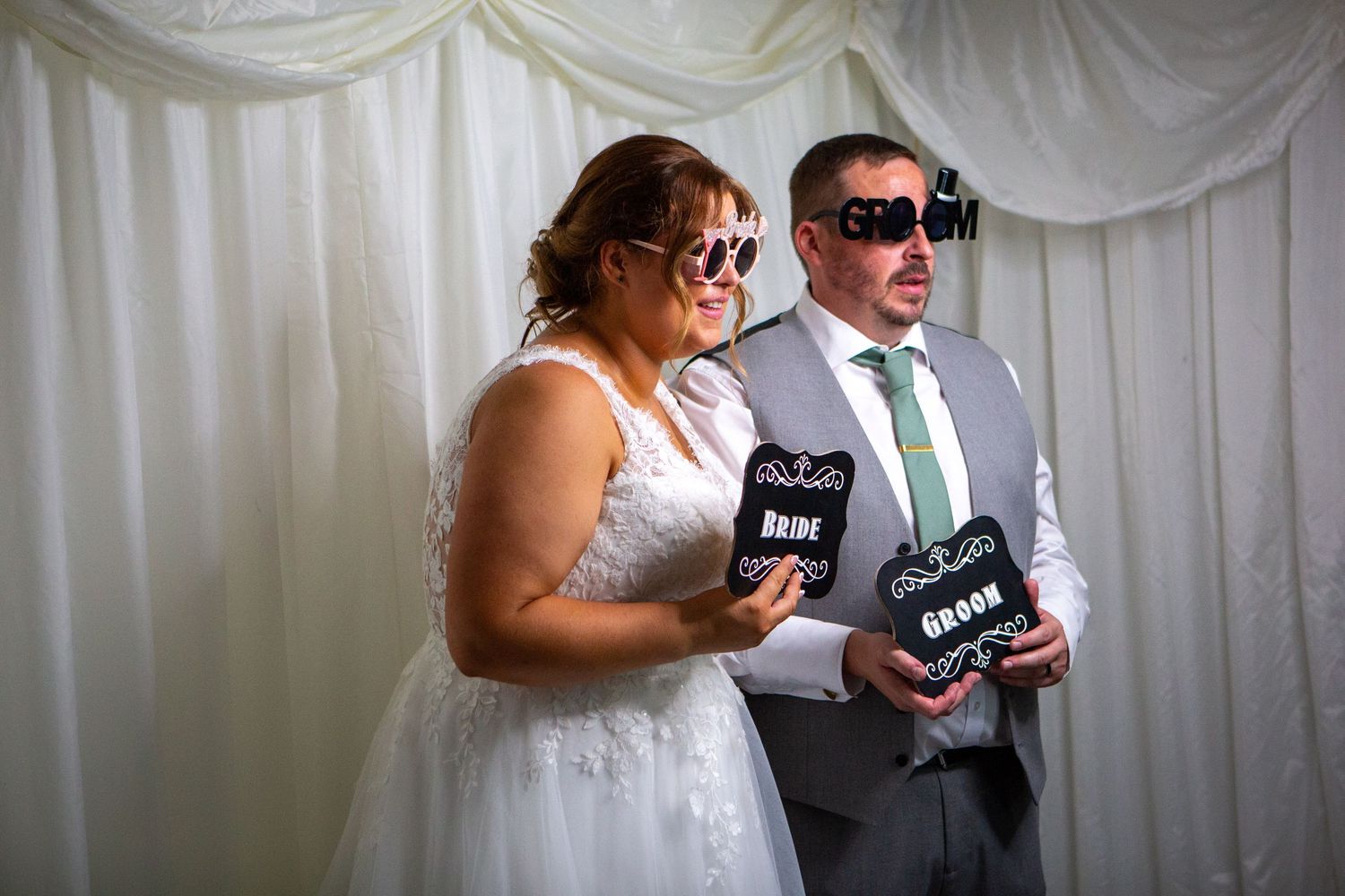 Bride and groom posing in a photo booth wearing fun novelty glasses and holding Bride and Groom signs at their wedding reception.