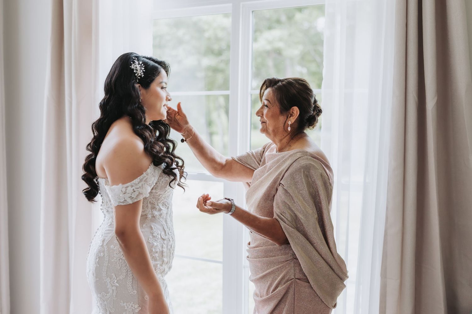 Candid photo of a bride with long dark curls and a jeweled hairpiece being helped by her mother in a light-filled bridal suite.