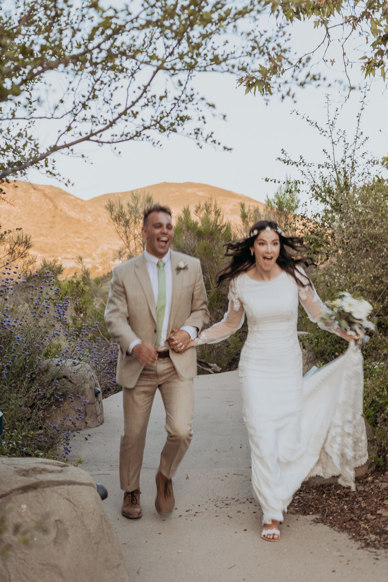 Joyful newlyweds in wedding attire run hand-in-hand on a sunlit path with desert hills in the background at Mission Trails Community Center.