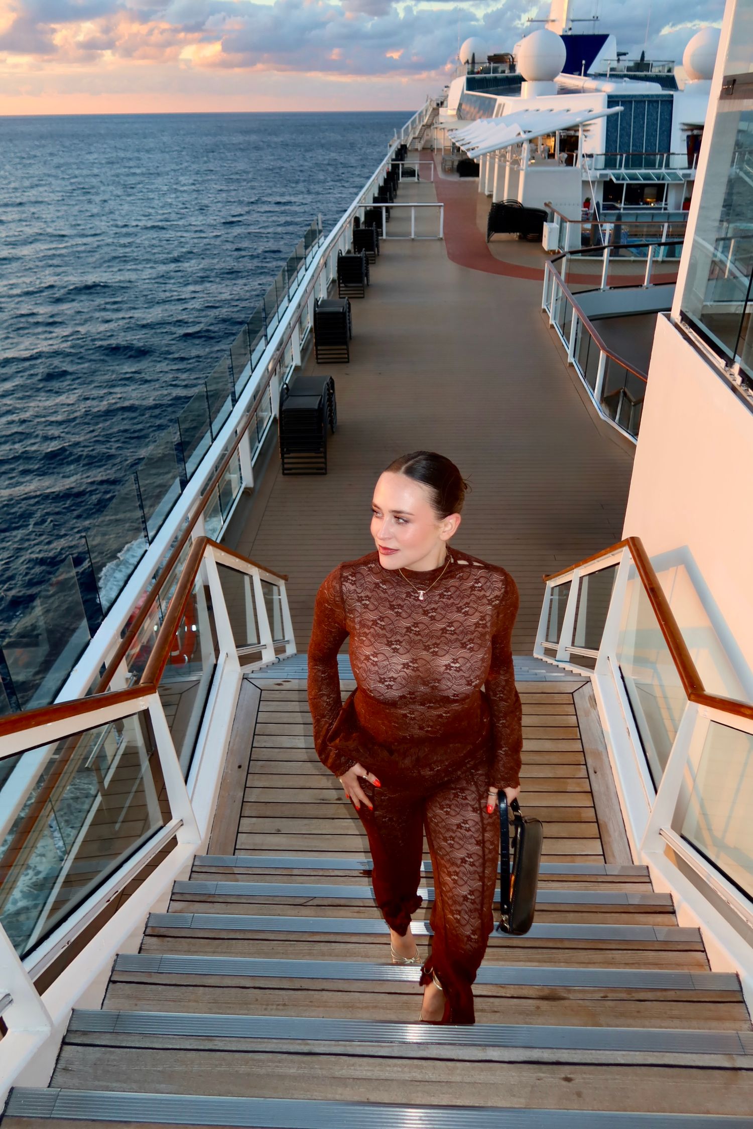 Woman in brown lace outfit ascending stairs on cruise ship deck at sunset with ocean views.