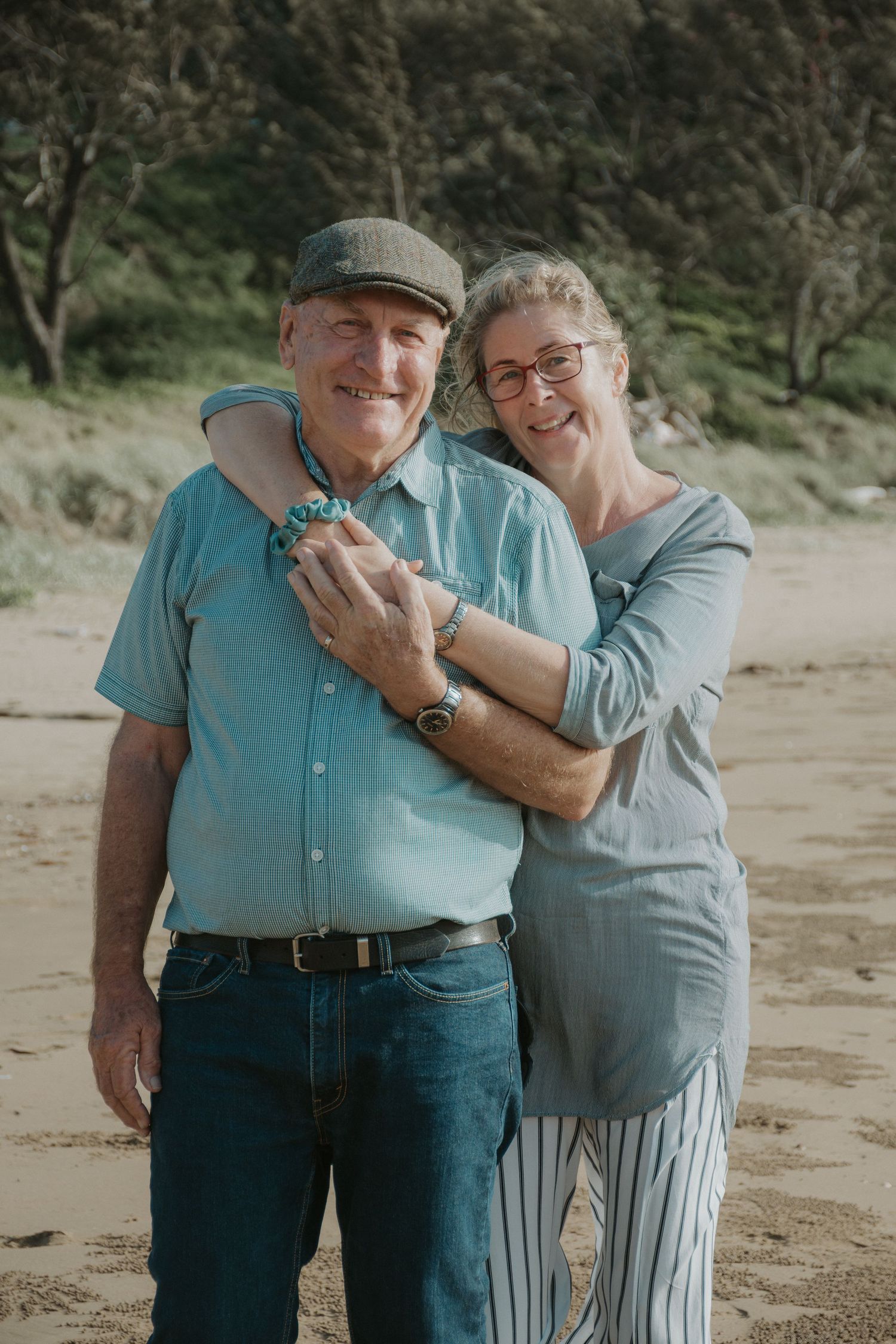 Happy older couple embracing on a sandy beach surrounded by coastal trees.