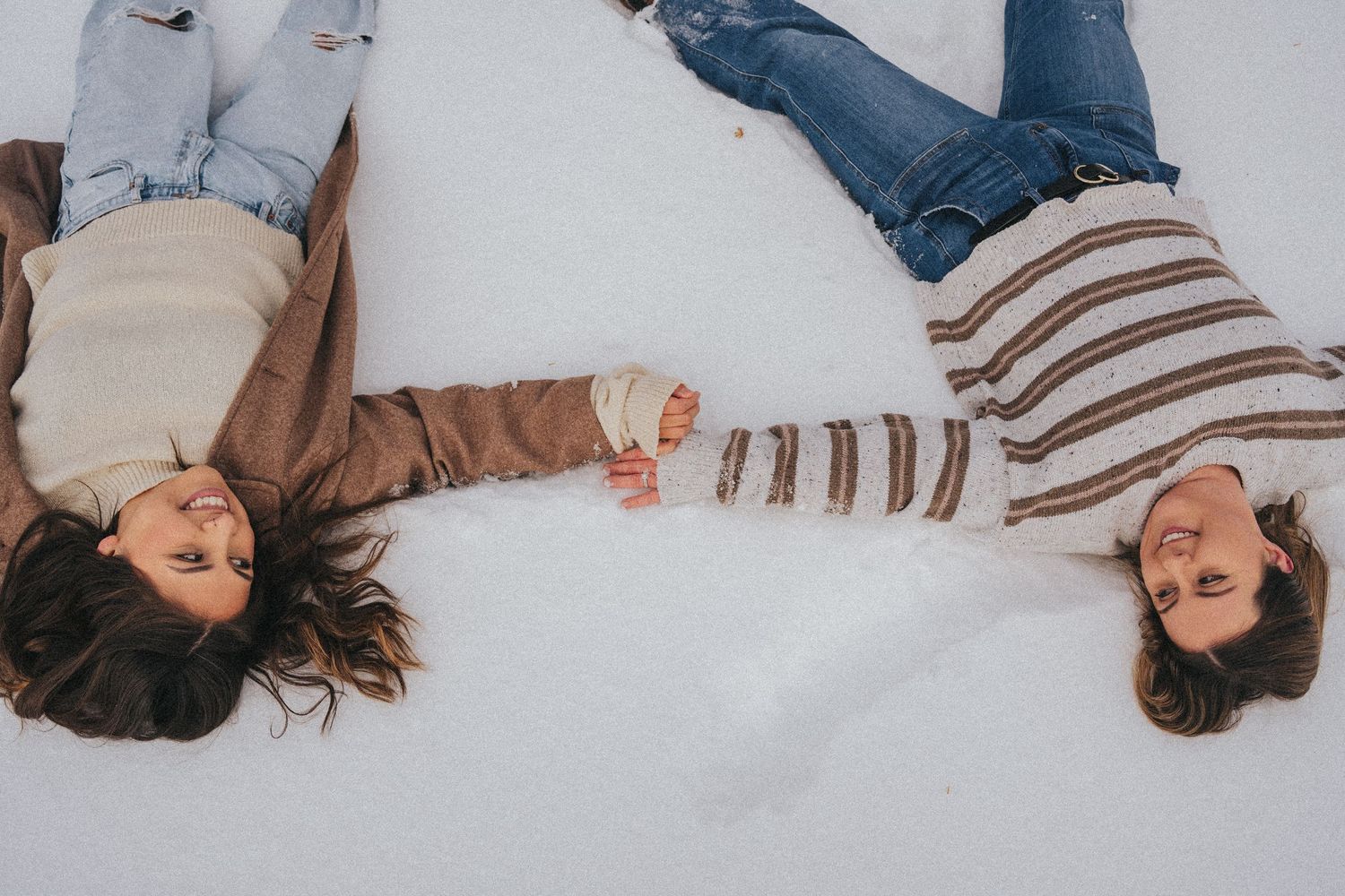 Two women lying in snow holding hands, wearing cozy winter outfits, smiling and enjoying a snowy day together.