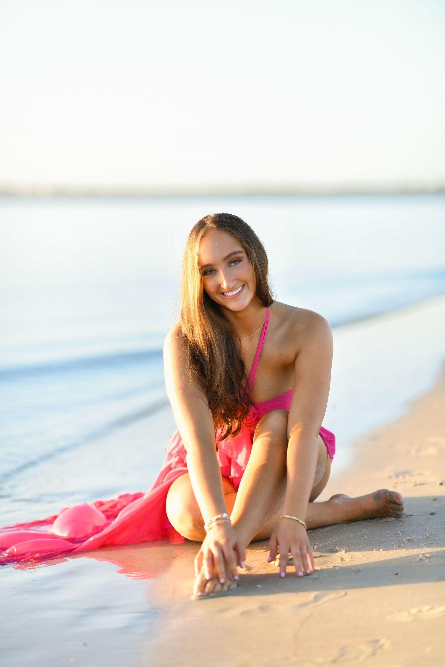 Smiling young woman in pink bikini sitting on sandy beach shore with calm blue water in the background.