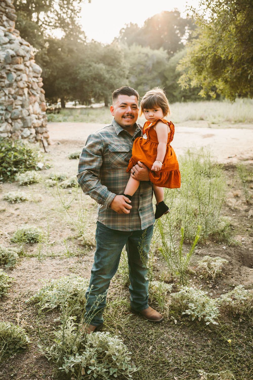 A man in a plaid shirt holds a toddler girl in an orange dress outdoors near stone walls and trees.