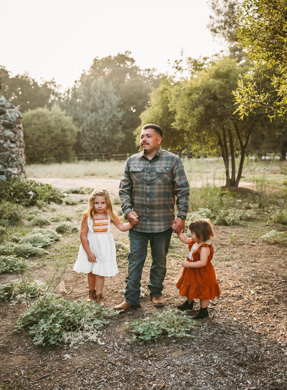 A father holds hands with two young daughters in a sunlit outdoor park setting surrounded by lush green trees.