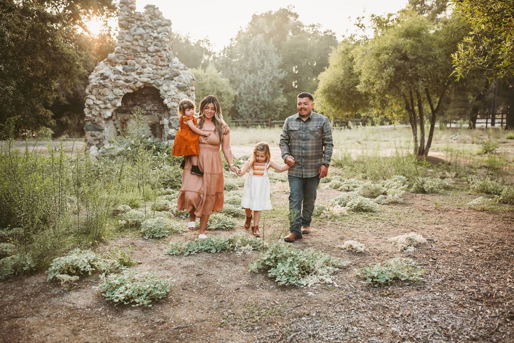 Family of four walking outdoors in a lush garden park with stone ruins and golden sunlight filtering through trees.