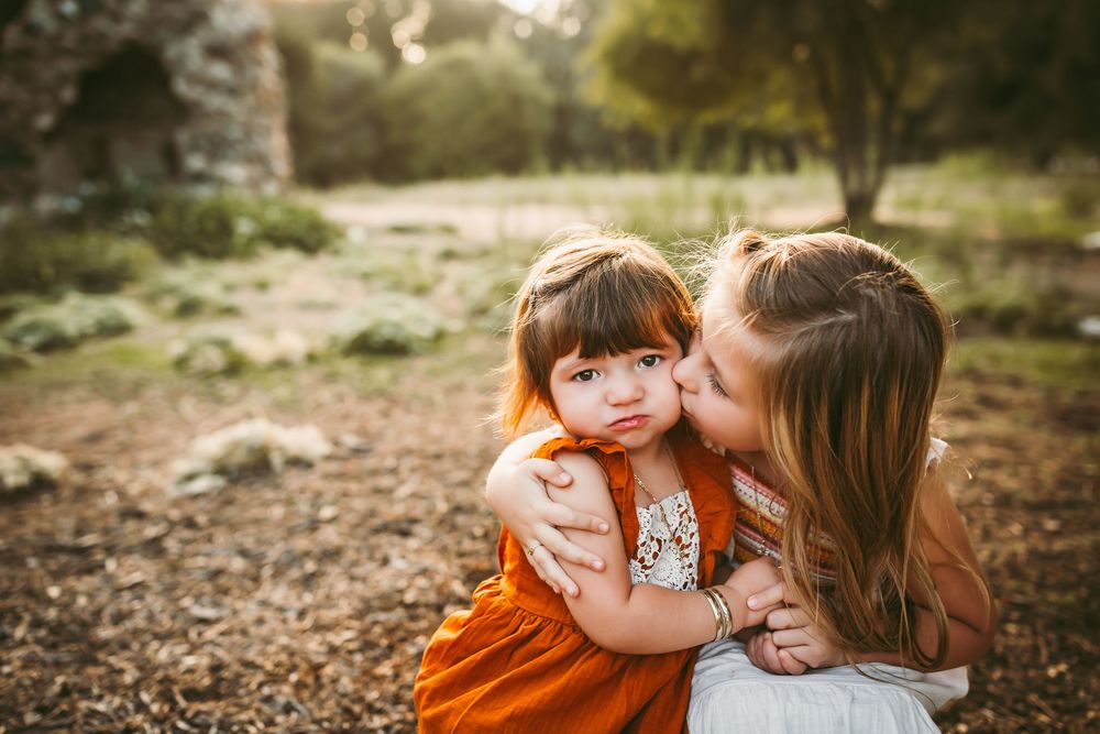 Older girl kissing younger toddler girl on cheek outdoors in a sunlit park setting.