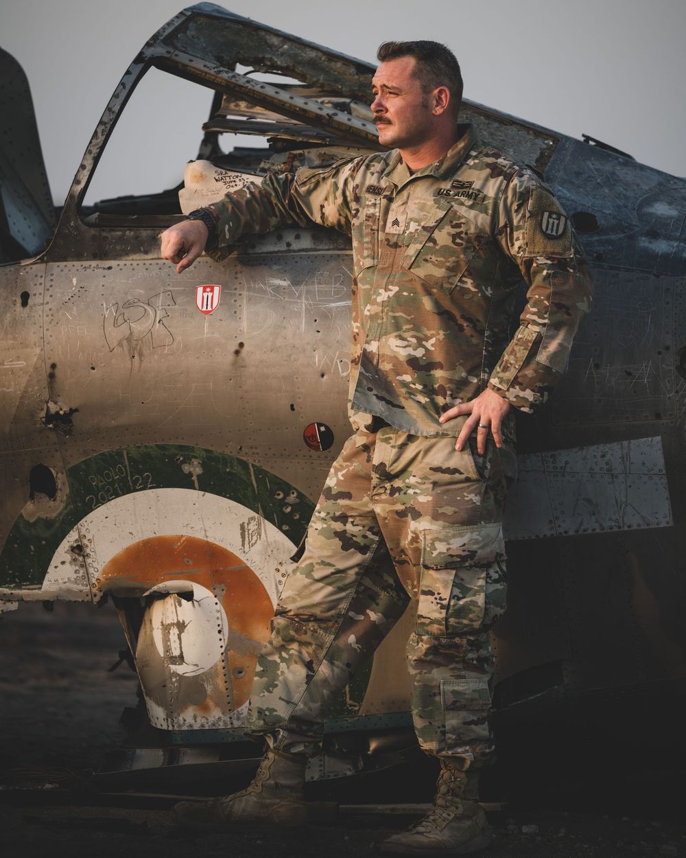 A soldier in camouflage uniform stands confidently beside a weathered military aircraft outdoors.