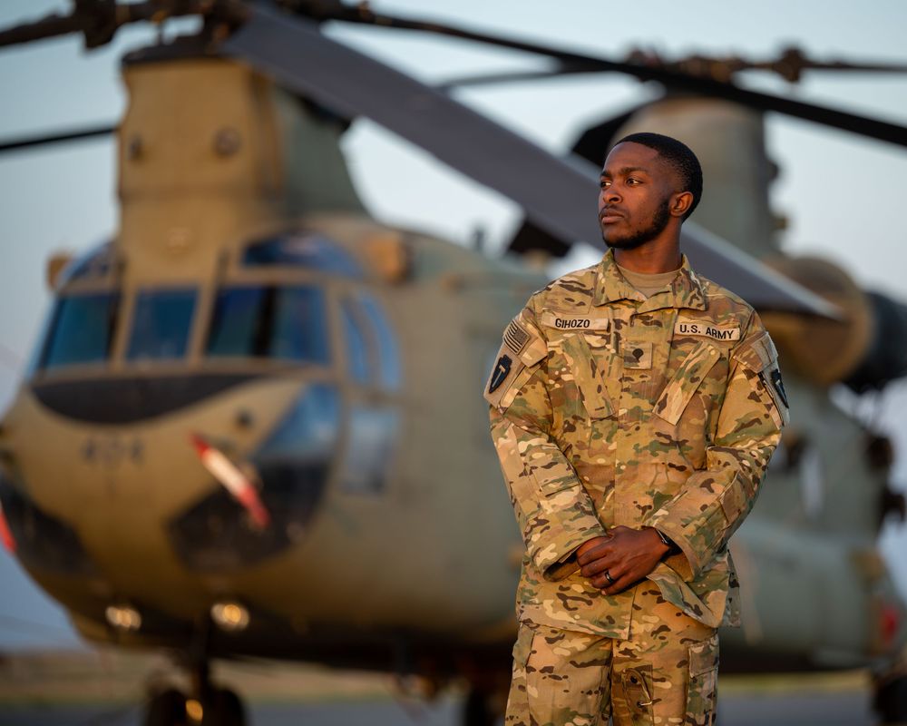 U.S. Army soldier in camouflage uniform stands confidently before a CH-47 Chinook helicopter at dusk.