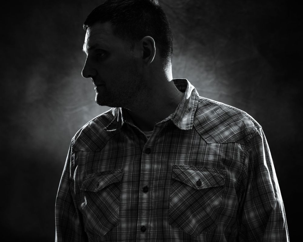 Black and white dramatic portrait of a man in a plaid shirt, shot in low-key lighting against a dark background.