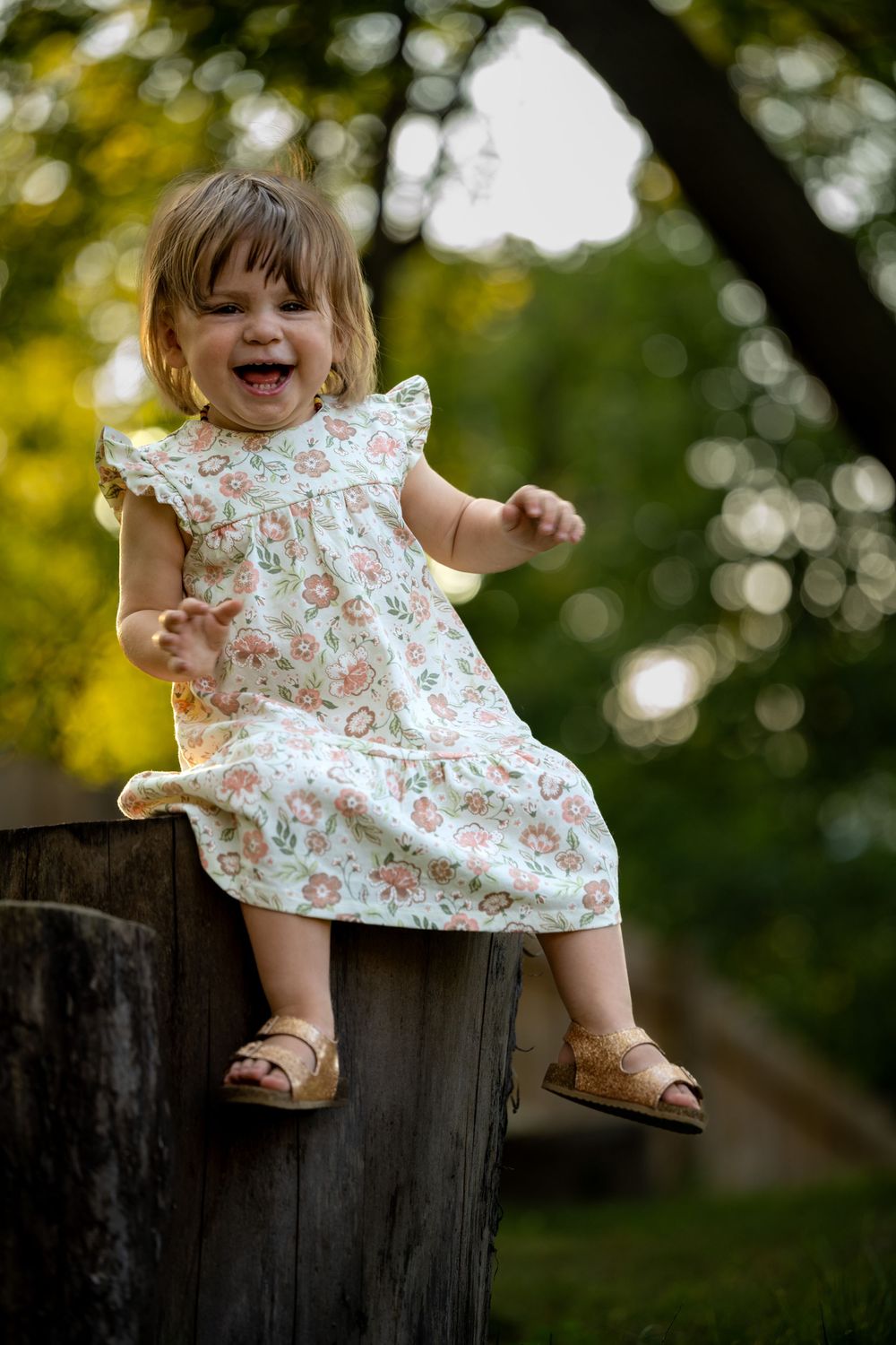 Laughing toddler girl in floral dress and sandals sitting on wooden stump outdoors in green park setting.