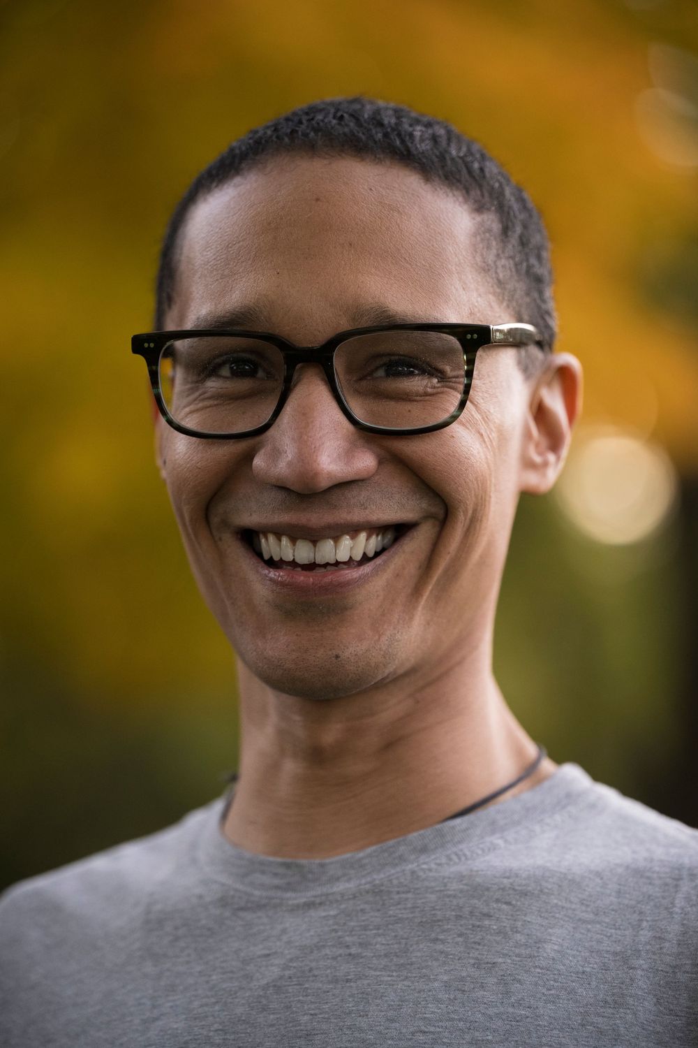 Smiling man with glasses and short hair wearing a gray t-shirt outdoors with autumn foliage background.
