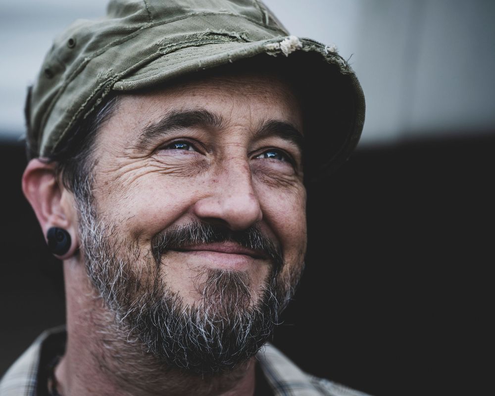 Smiling bearded man wearing a green cap and ear gauge, with blue eyes and a goatee, captured in a candid outdoor portrait.