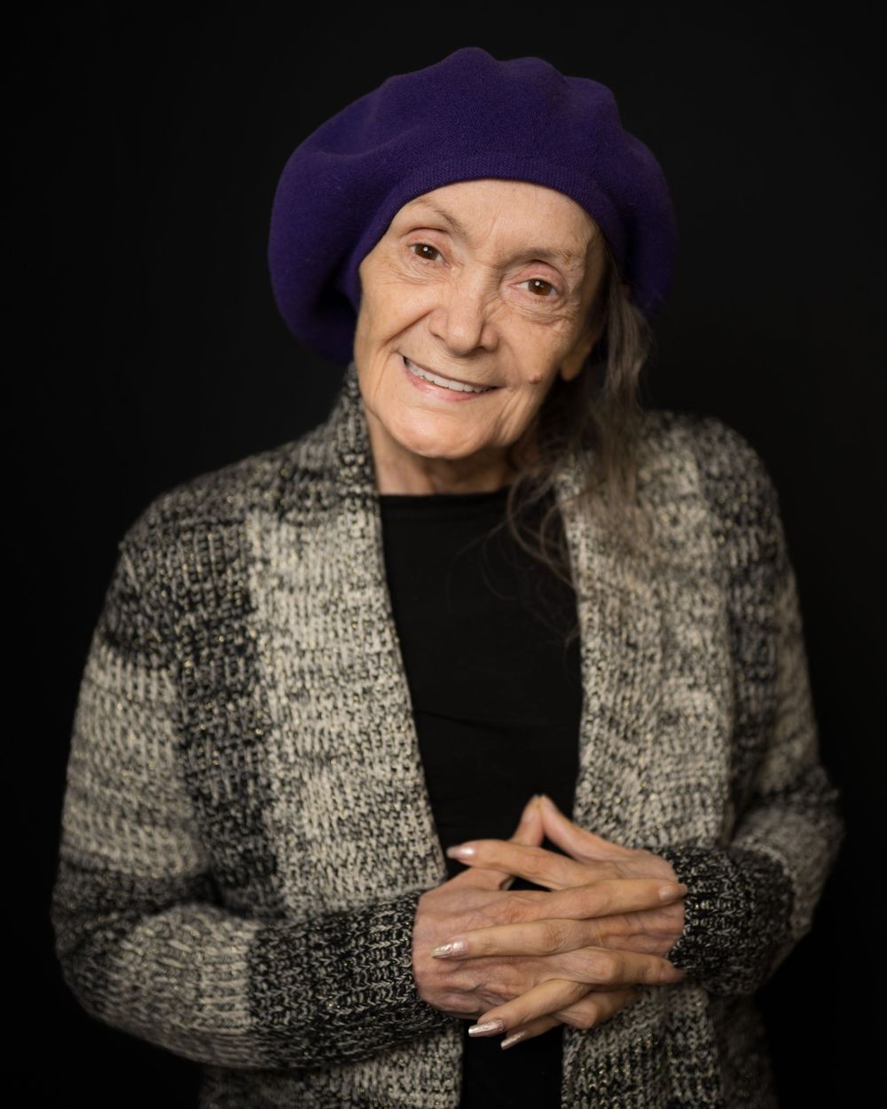 Elderly woman smiling warmly, wearing a purple beret and gray knit cardigan, posing against a dark background.