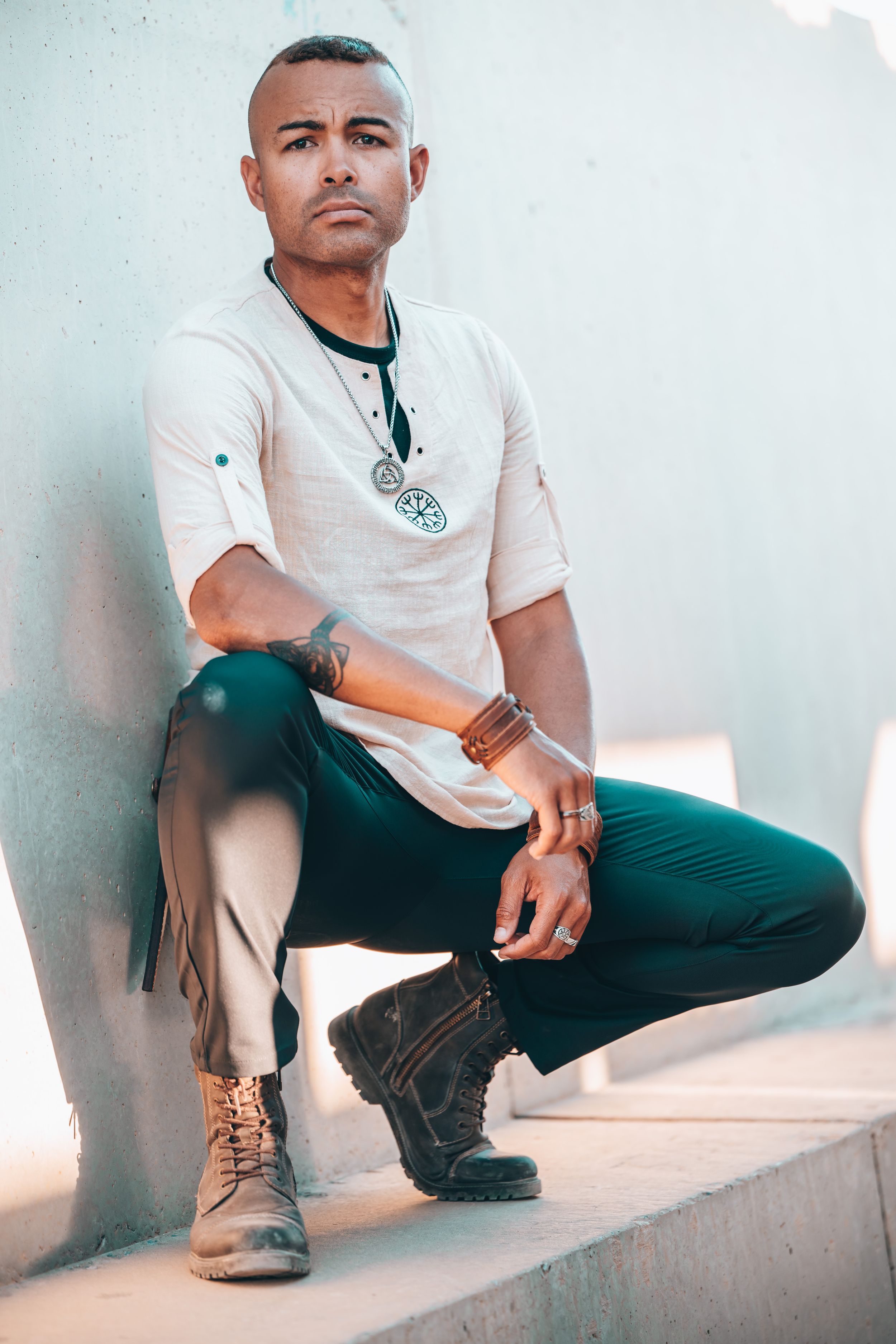 Man in white t-shirt, teal pants, and boots crouching on steps, wearing a pendant necklace and leather bracelets.